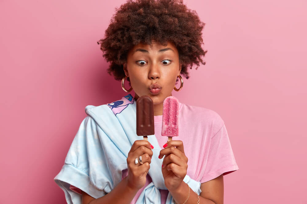 young woman in pink background holding two different flavoured popsicles